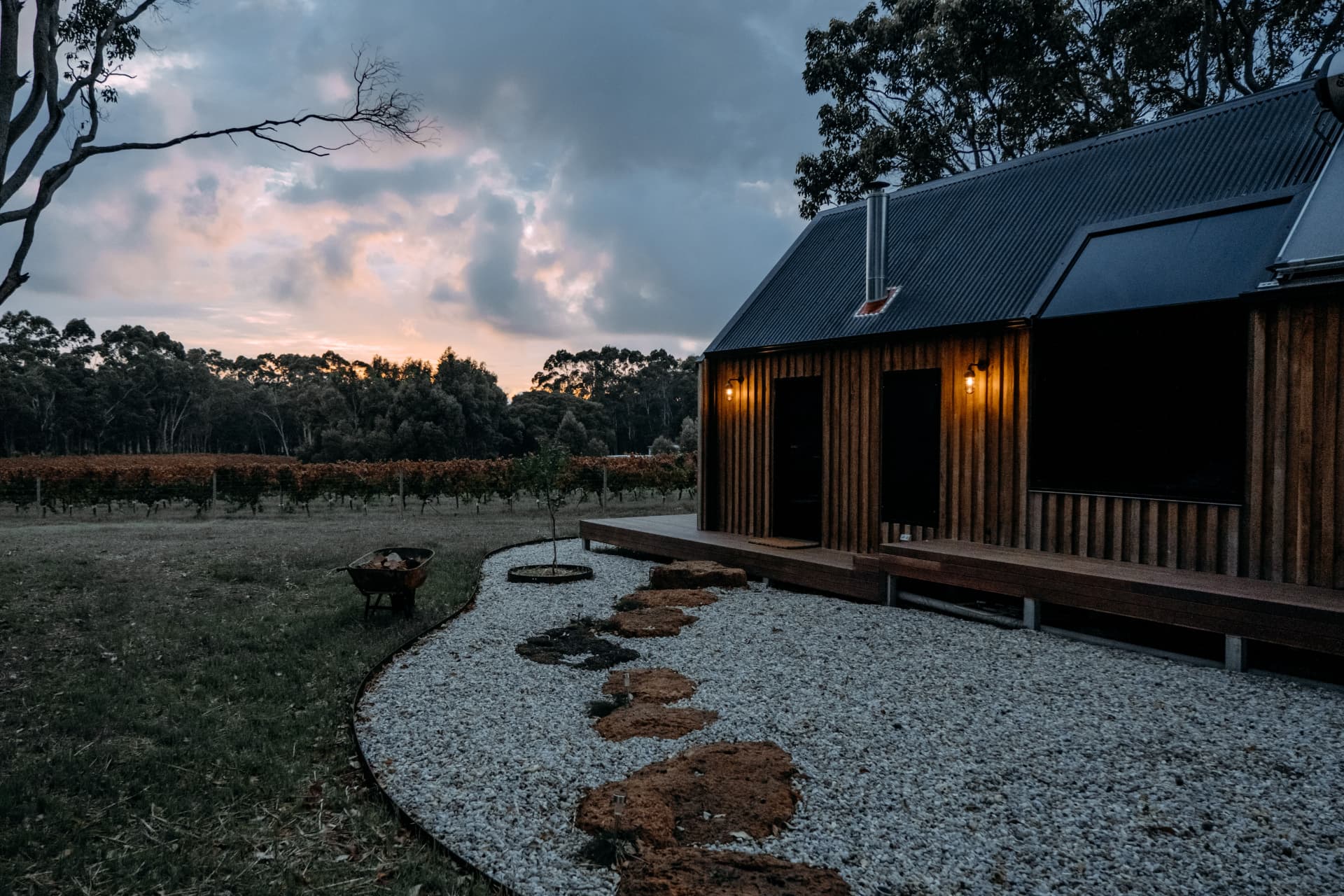 A timber cabin lit at dusk beside a vineyard and gum trees